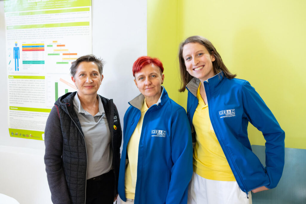 Drei Pflegerinnen in blauen GGZ-Jacken stehen vor einer grünen Wand und lächeln.