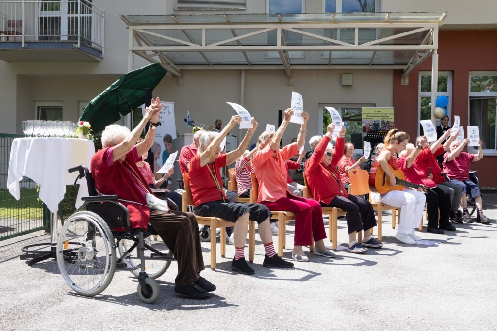 Senior:innen sitzen in einer Gruppe auf Stühlen und machen eine Gesangsvorstellung bei der 10 Jahres Feier des Memory Tageszentrums und Pflegewohnheims Aigner Rollett