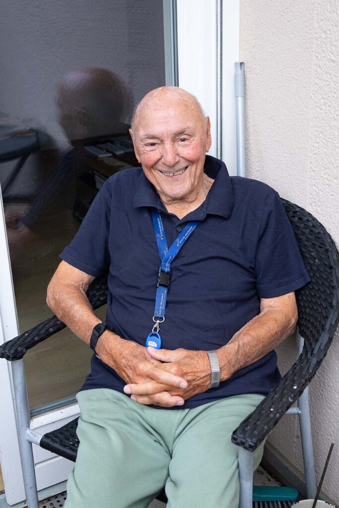 Senior sitzt auf dem Balkon seiner Wohnung auf einem Sessel und lächelt in die Kamera