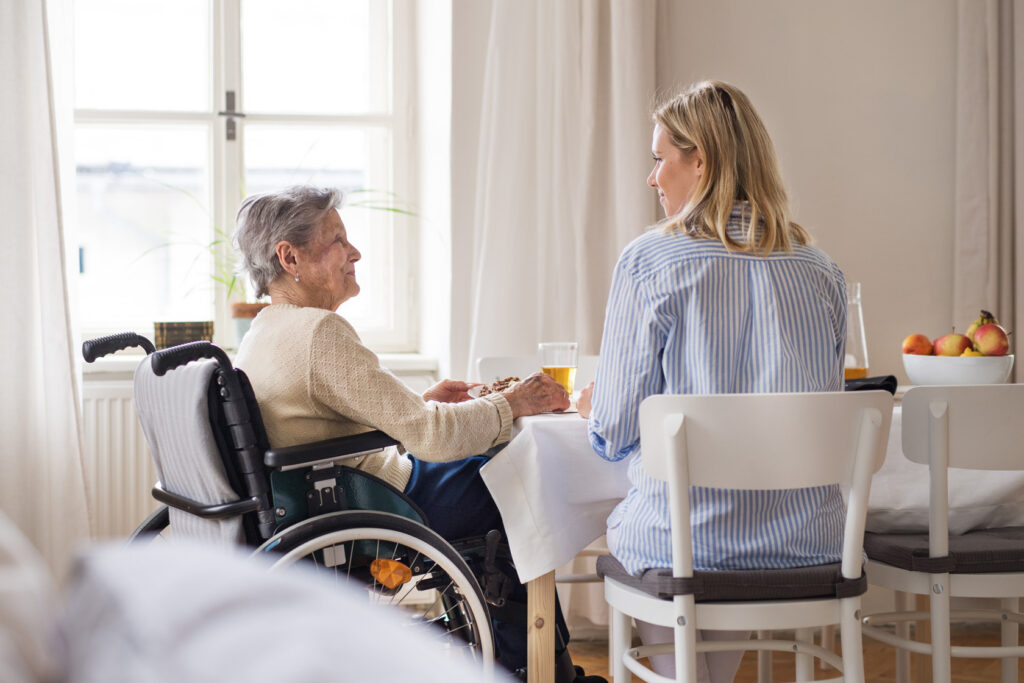 Pflegende Angehörige sitzt am Tisch mit pflegebedürftiger Person im Rollstuhl