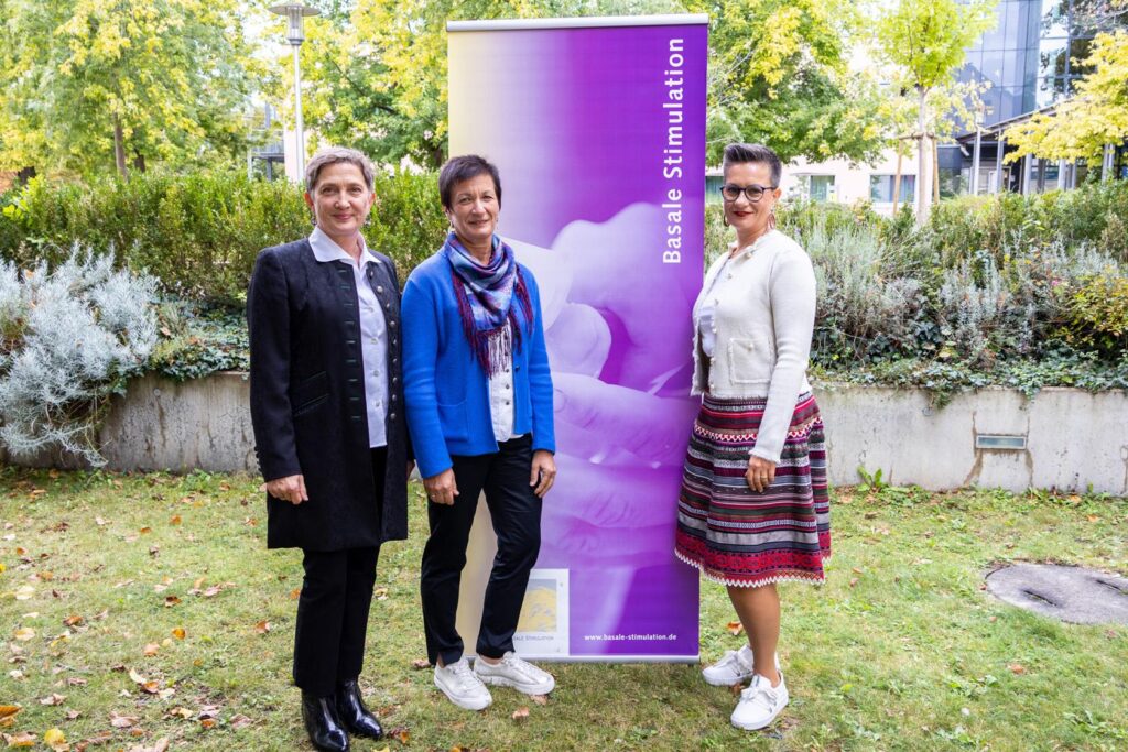 Die drei Kursleiter:innen posieren vor dem Rollup der Basalen Stimulation.