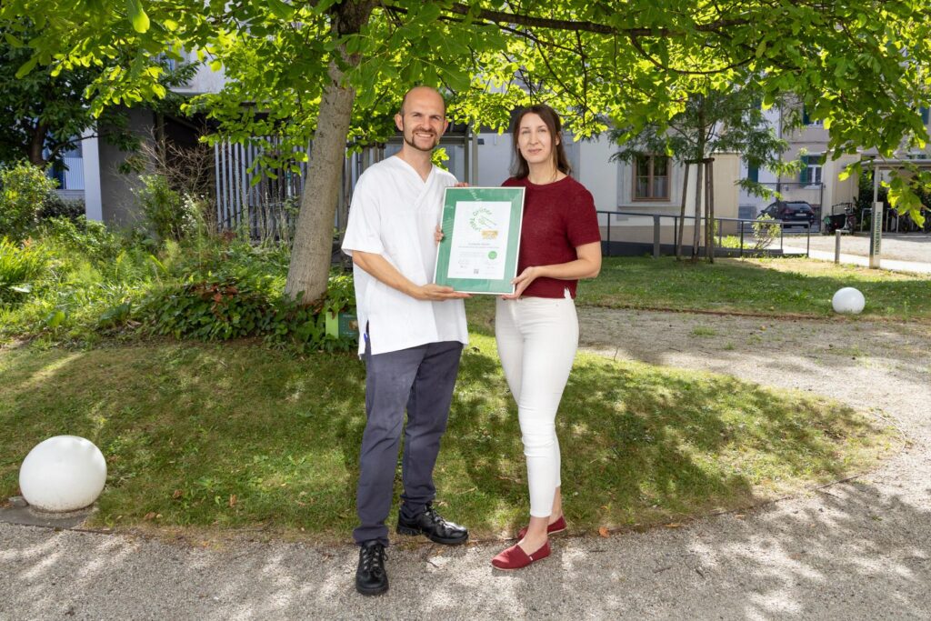 Zwei Personen stehen im Außenbereich der GGZ und halten die Auszeichnungsurkunde.
