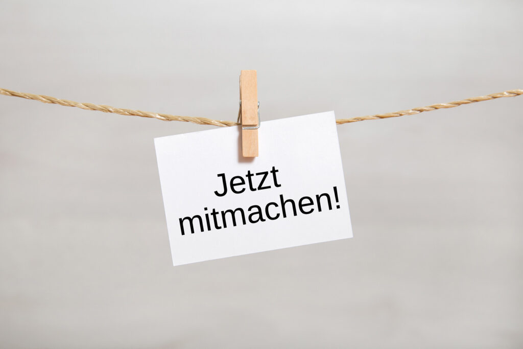 Ein an einer Schnur befestigter Zettel, der mit einer Wäscheklammer fixiert ist und die Aufschrift „Jetzt mitmachen!“ trägt.