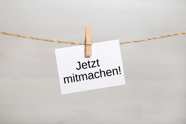 Ein an einer Schnur befestigter Zettel, der mit einer Wäscheklammer fixiert ist und die Aufschrift „Jetzt mitmachen!“ trägt.